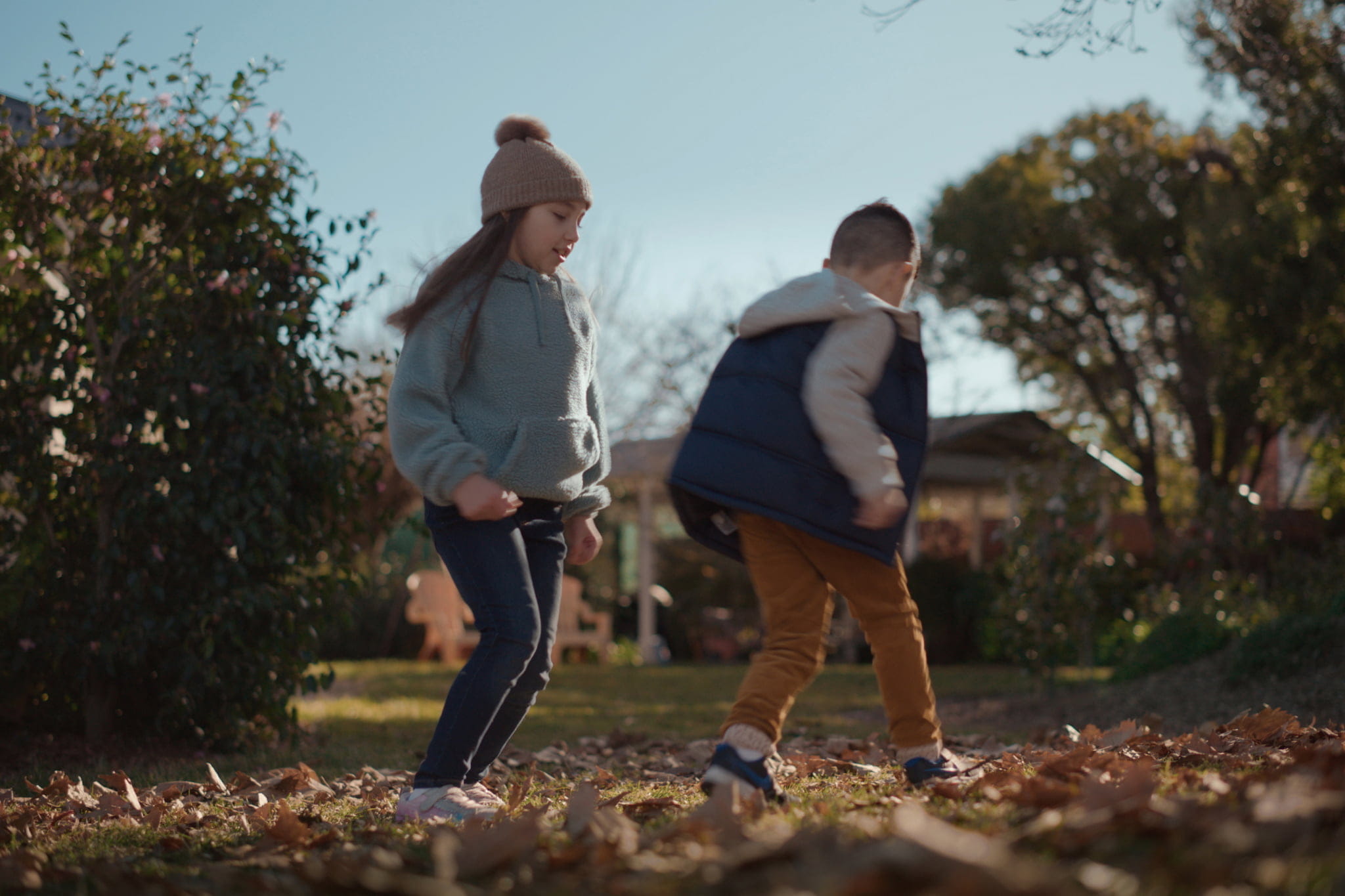 Two children wearing winter clothing playing in the fallen leaves