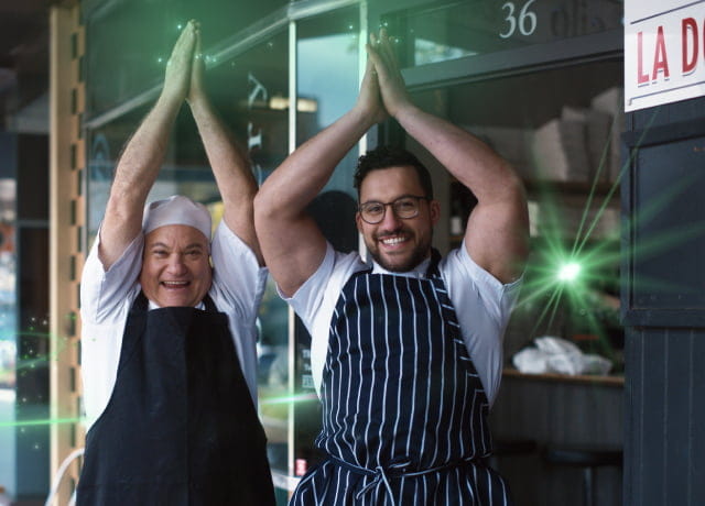 Two chefs standing outside their local restaurant celebrating with their arms in the air