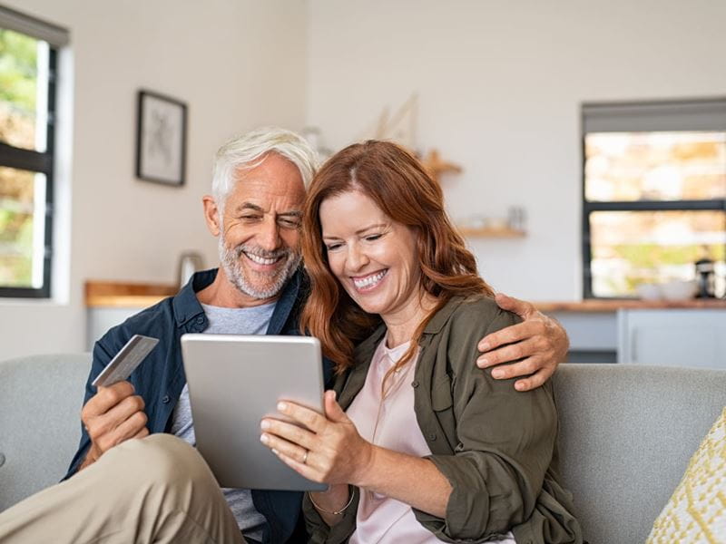 A couple with smiles on their faces, using a tablet while sitting on the lounge