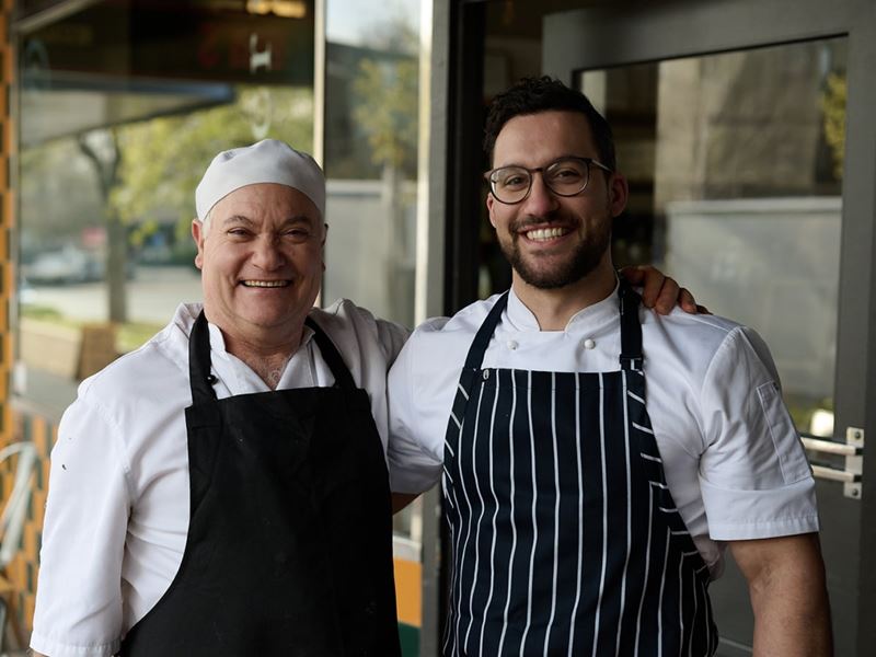 Two men wearing white shirts and aprons standing outside a local small business